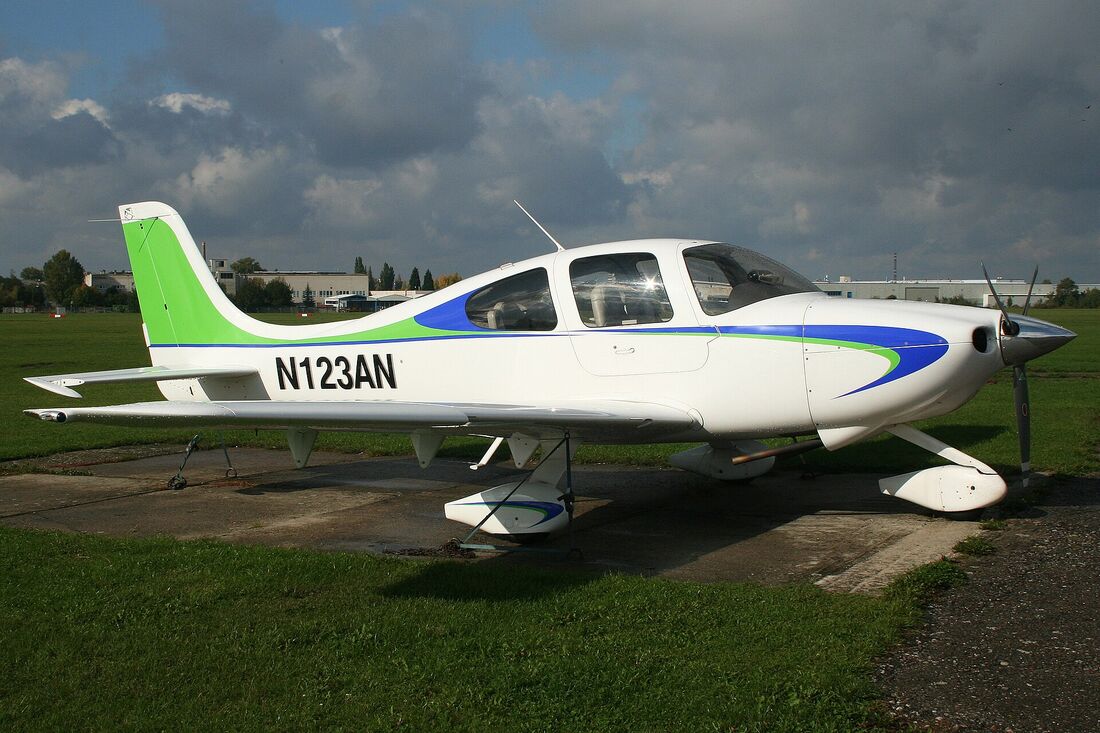 A white Cirrus SR20 with green and blue trim parked on an airfield, seen from the side