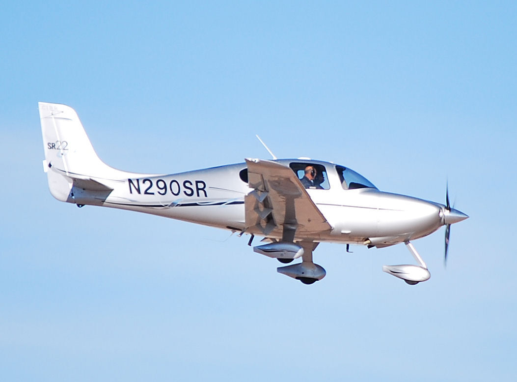 A white Cirrus SR22 flying against a clear blue sky, seen from the side, showing its low wing, swept tail fin and faired fixed landing gear